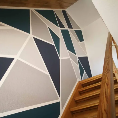 Kreative Wandgestaltung in einem Treppenhaus mit einem großflächigen, geometrischen Muster aus Dreiecken in Blau-, Grün- und Grautönen.